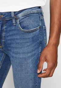 Jeans de mezclilla azul oscuro con corte slim, que cuentan con cinco bolsillos, costuras visibles y una textura sutil de desgaste en la tela.