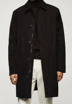 Homme portant un manteau noir boutonné et zippé par-dessus un pull à col roulé foncé et un pantalon blanc avec une ceinture noire, visible du cou jusqu'aux cuisses.