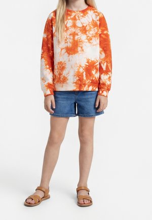 Barn iført orange og hvid tie-dye sweatshirt, blå denimshorts og tan sandaler, stående mod en ensfarvet hvid baggrund.
