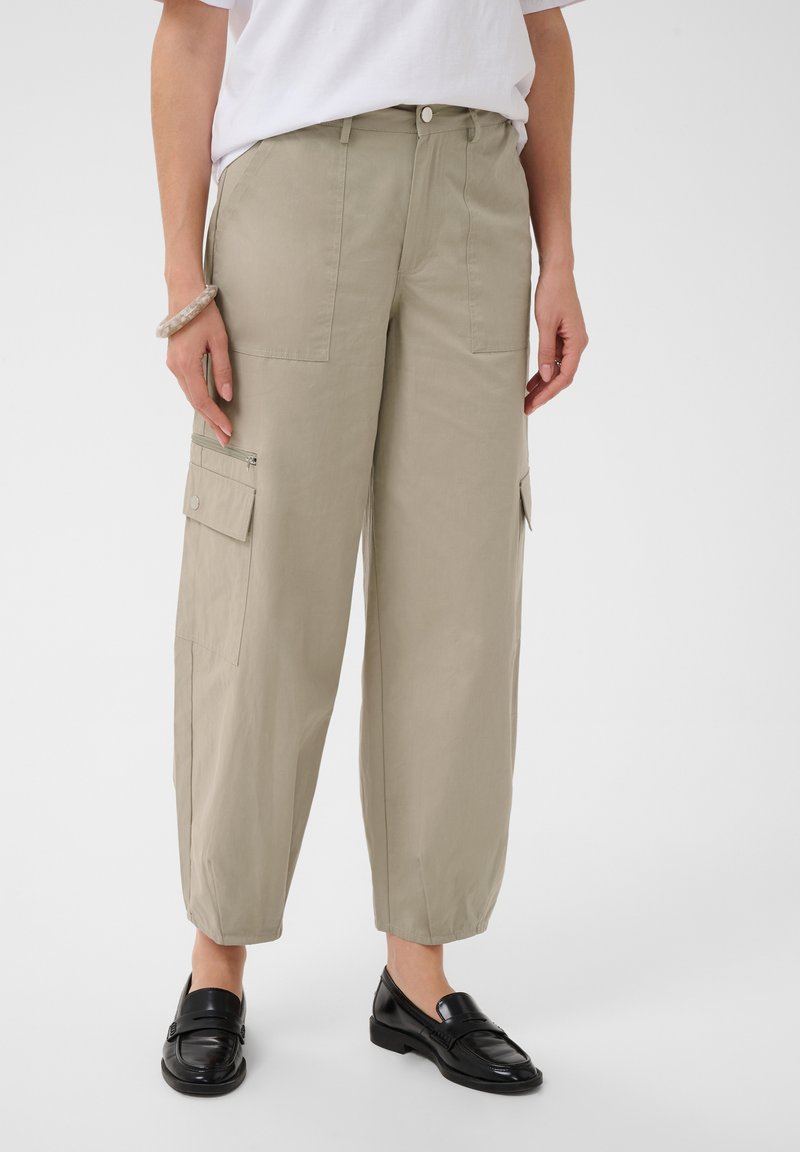 Personne portant un pantalon cargo beige clair à jambes larges avec poches à boutons et fermetures éclair, mocassins noirs, chemise blanche et un bracelet au poignet gauche.