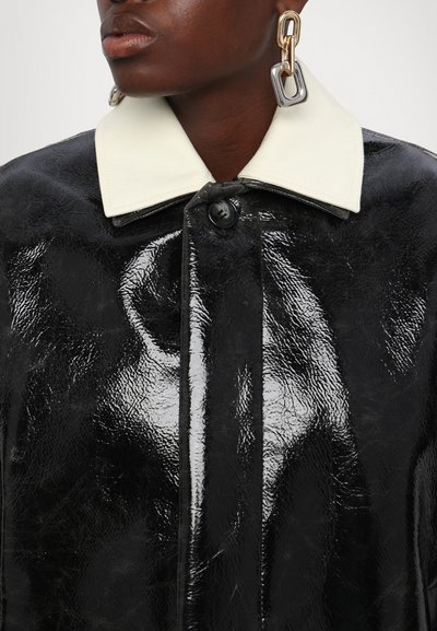 Cappotto lucido nero con finitura testurizzata, dotato di un colletto bianco a contrasto e chiusura frontale con bottoni. Orecchini di grandi dimensioni a catena come accenti.