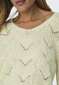 Maglione lavorato a maglia in morbido color crema, con un motivo a zigzag e dettagli in pizzo, e una scollatura ampia. Texture leggera.