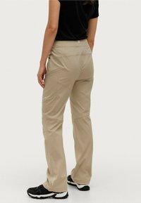 Beige wandelbroek met een gladde textuur, voorzien van zijzakken en een logo aan de achterkant. Gecombineerd met zwarte outdoorschoenen.