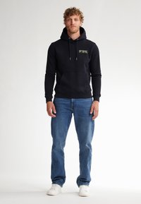 Sudadera negra con bolsillo frontal y detalle gráfico verde, combinada con jeans azules y zapatillas blancas. El tejido parece suave y casual.