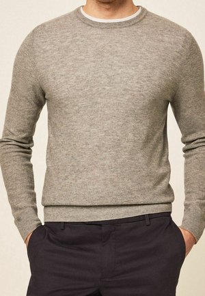 Mann trägt einen hellgrauen strukturierten Strickpullover mit gerippten Bündchen und Saum, kombiniert mit dunkler Hose, Hände in den Taschen.