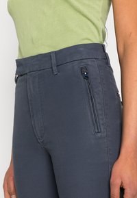 Calças cinza-escuras com uma textura suave, apresentando um bolso lateral com zíper e uma cintura plana. O tecido parece leve e flexível.