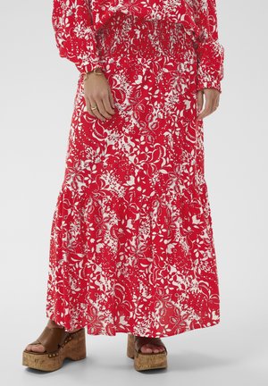 Žena má na sebe dlhú červenú a bielu kvetovanú sukňu s nariaseným pásom, hnedé platformové sandále, zlatý náramok a prstene na rukách.