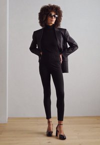 Combinaison ajustée noire avec ceinture élastique, superposée sous un blazer noir texturé. Le mannequin porte des lunettes de soleil et des talons hauts noirs.