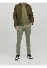 Olivgrön bomullsjacka över en olivfärgad skjorta, parat med ljusgröna byxor och svartvita sneakers, stående mot en grå bakgrund.