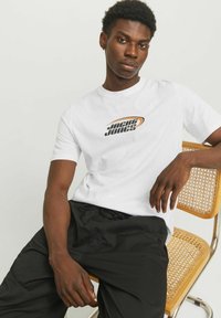 Witte katoenen t-shirt met 'Jack & Jones' logo in zwart en oranje. Gecombineerd met zwarte shorts. Model zit op een rieten stoel.