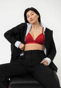 Punainen kolmionmuotoinen bralette, jossa on säädettävät olkaimet, kerrostettuna mustan muotisuunnittelijan bleiserin ja raidallisen paidan alle. Mustat korkeavyötäröiset housut näkyvissä.