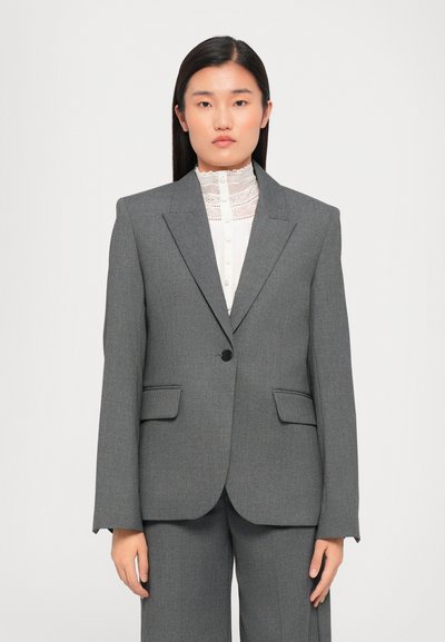 Grauer maßgeschneiderter Blazer mit einem einzigen Knopf, angedeuteten Revers und zwei Fronttaschen. Getragen über einer weißen Bluse mit hohem Kragen und Spitzendetails.