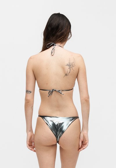 Γυναίκα με μακριά σκούρα μαλλιά που φοράει ασημί μεταλλικό μπικίνι, δείχνοντας την πλάτη με τατουάζ λουλουδιών και ματιών στα χέρια και τον ώμο.