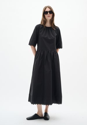 Femme portant une robe midi noire oversize avec des bords festonnés, des chaussures noires slip-on et de grandes lunettes de soleil foncées sur fond blanc.