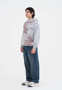 Felpa grigia chiara con grafica di un drago rosso, pantaloni larghi in denim blu e sneakers bianche con accenti neri e un design a motivo.