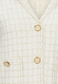 Blazer en tissu texturé crème avec un motif pied-de-poule subtil et trois boutons ronds en bois et or sur le devant.