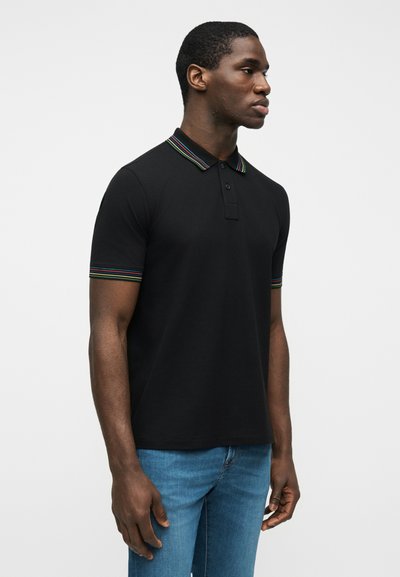 Άνδρας που φοράει μαύρο πόλο μπλουζάκι με κοντό μανίκι και πολύχρωμες ρίγες στο γιακά και στα άκρα των μανικιών, συνδυασμένο με μπλε τζιν, στέκεται πλαγίως.