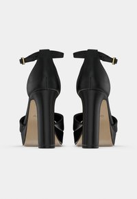 Mustat korkokengät, jotka on valmistettu sileästä nahasta, paksulla korolla, avoimella kärjellä ja nilkan ympärille kiertyvällä hihnalla, jossa on kultaiset metalliosat.