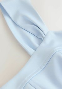 Tissu bleu clair avec un détail de nœud torsadé et des coutures visibles sur un matériau lisse et doux, présenté sur un fond neutre.