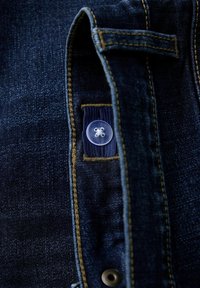Denim tkanina tamnoplave boje s zlatnim šavovima; sadrži prozirni plastični gumb i tkanu oznaku. Krupni plan detalja odjeće.