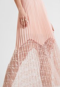 Robe plissée rose avec une superposition en dentelle au niveau de l'ourlet. Présente une texture douce et un design fluide, mettant en valeur des motifs de dentelle intriqués.