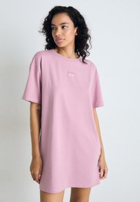 Kvinne med krøllete mørkt hår iført en løs rosa oversized T-skjorte-kjole med en liten "HUGO"-logo på brystet, stående mot en enkel hvit vegg.