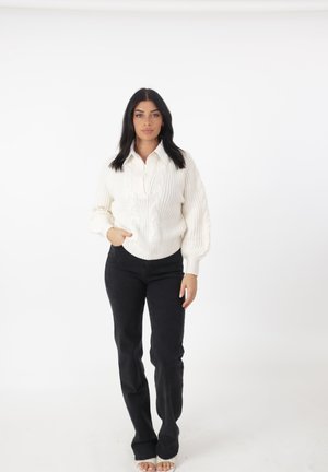 Witte kabelgebreide trui met een V-hals en geribbelde zoom, gecombineerd met zwarte flared jeans. Het model staat tegen een witte achtergrond.