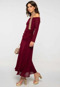 Robe bordeaux à épaules dénudées avec des détails froncés, manches longues, jupe fluide et une écharpe beige clair. Sandales à plateforme dorées et un petit sac.