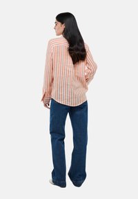 Chemise à rayures orange et blanche à manches longues avec un ourlet arrondi, portée avec un jean bleu taille haute. Tissu lisse avec une coupe décontractée.