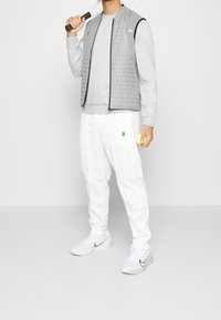 Grå vadderad väst över en grå sweatshirt, vita avsmalnande byxor och vita sneakers. Håller en tennisracket och en boll. Enkel design.