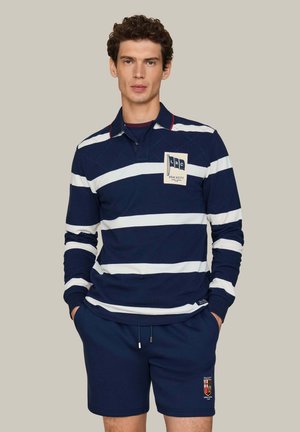 Hombre con camisa polo de manga larga a rayas azul marino y blanca y pantalones cortos azul marino con parches de marca, de pie con las manos en los bolsillos.