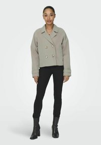 Chaqueta corta de corte cruzado en gris claro, con grandes botones y puños enrollados, combinada con jeans skinny negros y botines de tacón puntiagudos en negro.