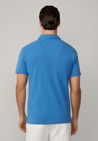 Kortärmad polo skjorta i klarblå bomull med en platt krage, sidokilningar vid fållen och en slät yta. Bakvy visas.