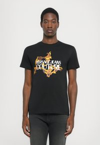 Črna bombažna majica s kratkimi rokavi, okroglo izrezana, z barvitim cvetličnim grafičnim vzorcem in besedilom "VERSACE JEANS COUTURE" v beli barvi.