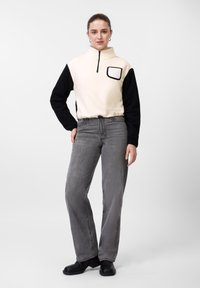 Φλις πουλόβερ με εκρού μπροστινό μέρος και μαύρα μανίκια, με γιακά με ημί-φερμουάρ, τσέπη στο στήθος· συνδυασμένο με ψηλόμεσα γκρι τζιν και μαύρα παπούτσια.