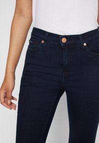 Mörkblå denimjeans med en figursydd design, utrustad med en kopparknapp vid midjan, dekorativ söm och femficksdesign.