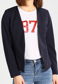 Kvinna i en marinblå öppen blazer över en vit T-shirt med röd "973"-tryck och blå jeans, stående mot en enkel bakgrund.