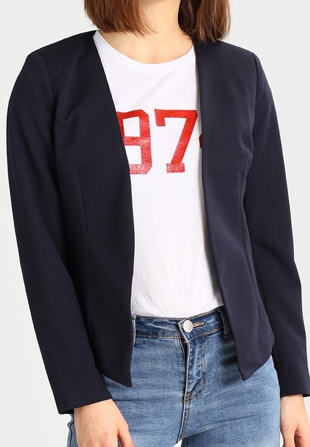 Kvinna i en marinblå öppen blazer över en vit T-shirt med röd "973"-tryck och blå jeans, stående mot en enkel bakgrund.