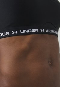 Juodas sportinis liemenukas su lygia tekstūra ir plačia elastine juosta, ant kurios baltais rašmenimis užrašyta „UNDER ARMOUR“. Švarus, minimalus dizainas.