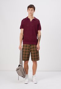 Bordeaux tekstureret strik polo skjorte og brune ternede shorts. Modellen holder en grå crossbody taske og har hvide sneakers på. Neutral baggrund.