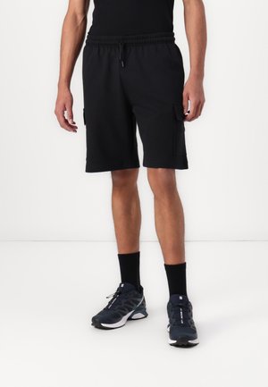 Homme portant un short cargo noir, des chaussettes noires et des chaussures de course noires et blanches, debout devant un fond blanc.
