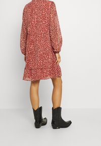 Červené šaty s vzorem, nafouknutými rukávy a volánkovým lemem, doplněné černými kovbojskými botami. Šaty jsou vyrobeny z lehkého, průsvitného materiálu.