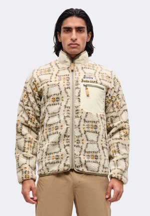 Άνδρας φοράει μπεζ fleece μπουφάν με σχέδιο, με φερμουάρ μπροστά και τσέπη στο στήθος με φερμουάρ, συνδυασμένο με ανοιχτά καφέ παντελόνια.