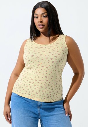 Donna con capelli lunghi e lisci che indossa una canotta gialla senza maniche con piccoli fiori rosa e jeans blu, in piedi davanti a uno sfondo bianco.