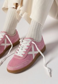 Baskets en suède rose avec des accents blancs, lacets plats blancs et semelle en caoutchouc brun texturé. Les chaussettes sont crème à côtes, partiellement visibles.