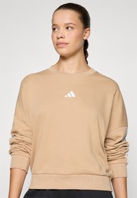 Beige bomullstrøye med rund hals, avslappet passform, og hvit Adidas-logo sentrert på brystet. Ribbet mansjetter og nederkant.