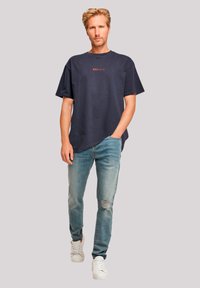 Marineblauwe katoenen t-shirt met korte mouwen en ronde hals; met rode tekst. Gecombineerd met lichtblauwe versleten spijkerbroek en witte sneakers.