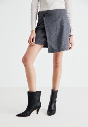 Person iført en grå slå-om nederdel med knap og sorte midtkalv stiletstøvler, stående mod en ensfarvet lys baggrund.