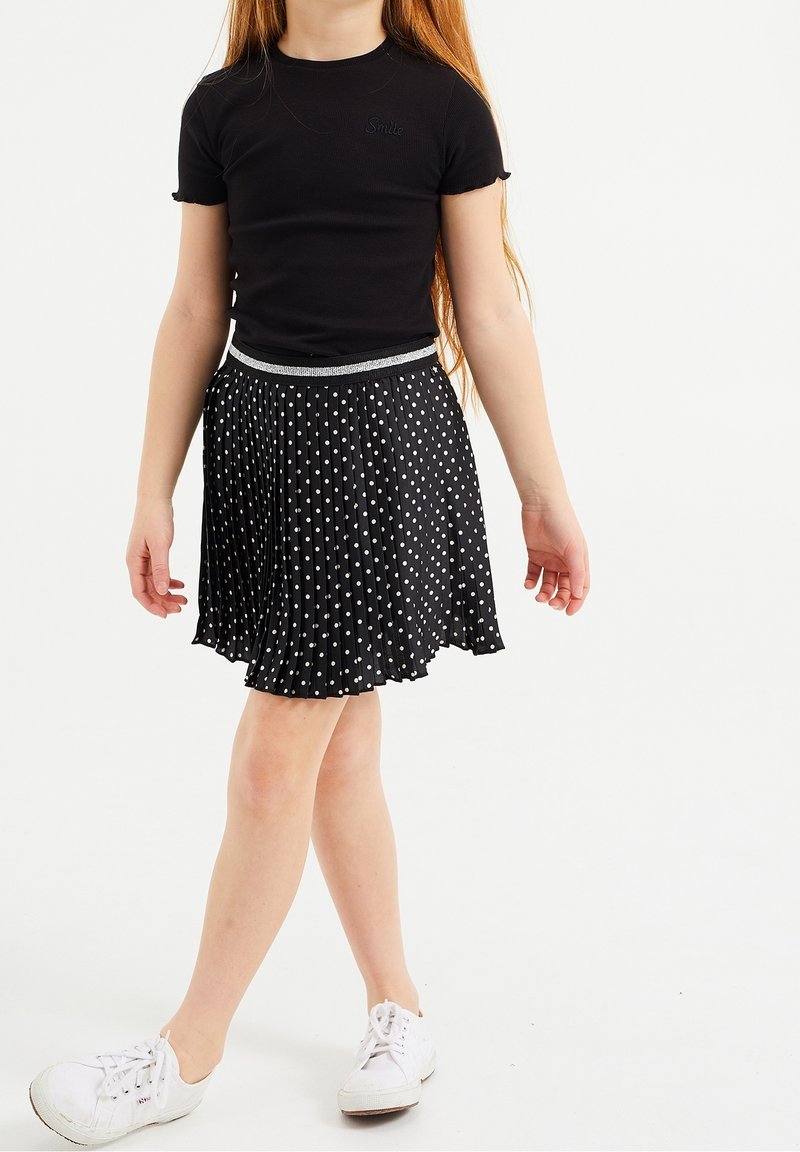 Fille portant un haut noir à manches courtes, une jupe plissée noire à pois blancs, et des baskets blanches, marchant avec les jambes croisées.