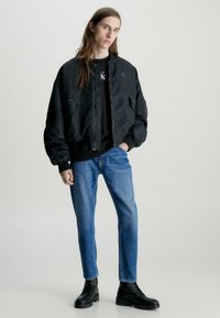 Chaqueta bomber negra con puños y dobladillo acanalados, combinada con una camisa oscura, jeans azules y botas de combate negras, sobre un fondo liso.